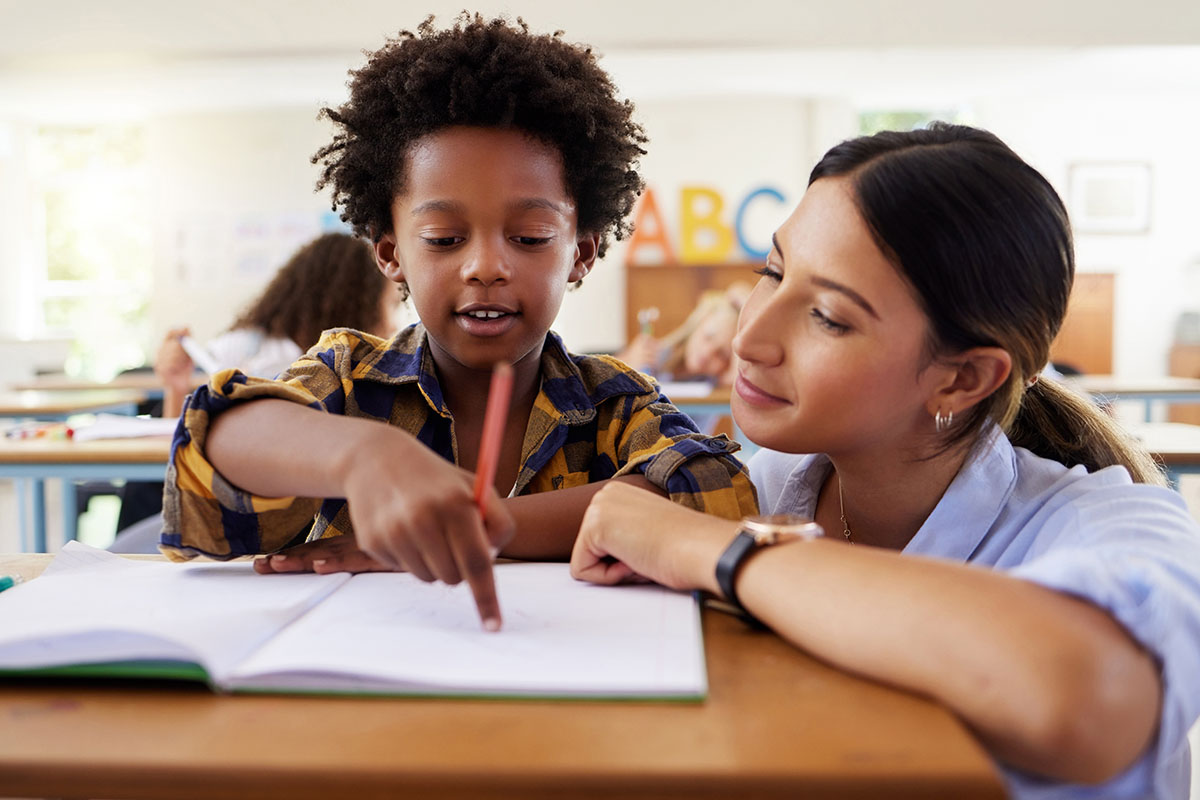 teacher, learning and helping black kid in classroom for knowledge, studying or assessment. question, development and boy or student with woman for education pointing in notebook in kindergarten.